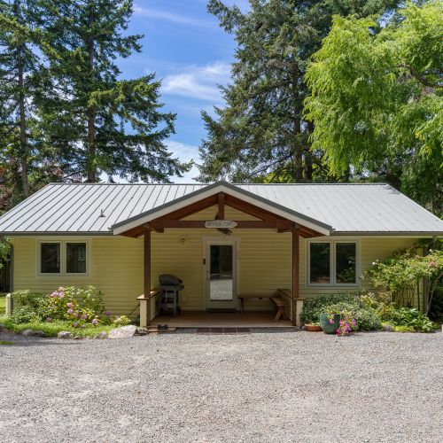 A charming cottage surrounded by mature trees, featuring a welcoming covered porch, beautiful landscaping, and convenient on-site parking.