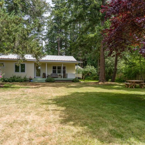Relax in your own private backyard retreat, featuring a spacious lawn, covered porch, and picnic table surrounded by towering evergreens and lush landscaping. It's the perfect place for outdoor dining, lawn games, or simply unwinding in a peaceful