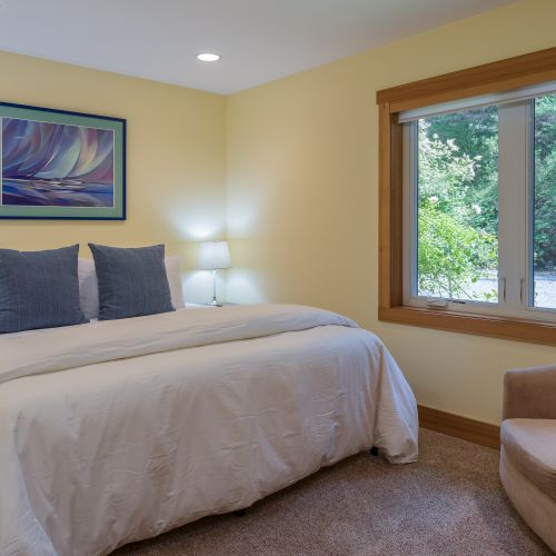 Relax in this bright queen bedroom featuring soft bedding, bedside lighting, a comfortable reading chair, and a large window overlooking the peaceful wooded surroundings.