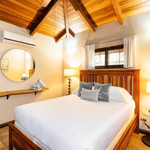 Cozy queen bedroom — vaulted exposed beams, hardwood slat headboard, warm bedside lamps.
