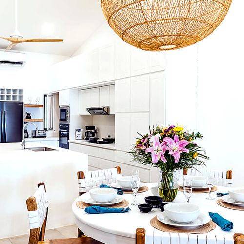 Round dining table set with place settings and fresh flowers, beneath vaulted ceilings and rattan pendant lighting, adjacent to the kitchen