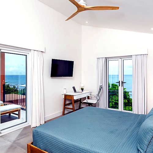 Primary bedroom featuring a king bed, desk, flat-screen TV, and French doors leading to a private ocean-view terrace.