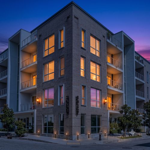 Modern multi-story building w/ private balconies & lush greenery out front.