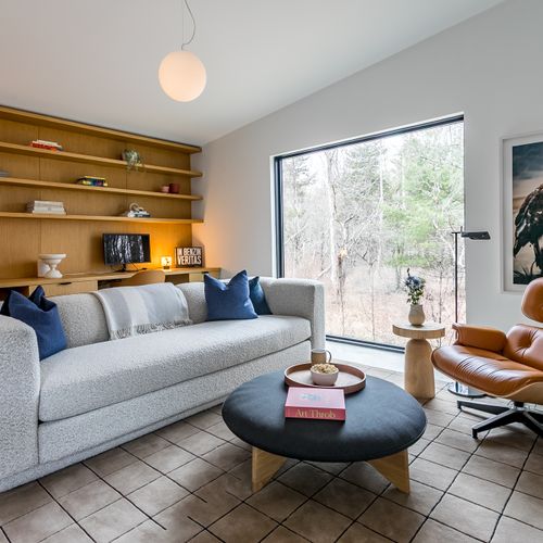 A bright, book-lined living area with a built-in oak shelving nook, plush sofa, and a leather lounge chair framing the forest through floor-to-ceiling glass.