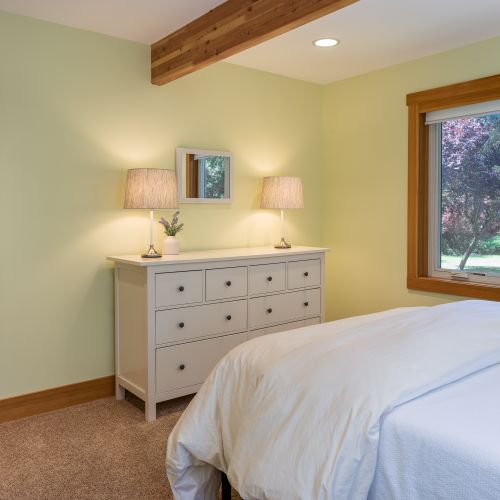 Natural light fills the room through a large picture window, while a spacious dresser and soft bedside lighting create a comfortable, relaxing retreat.