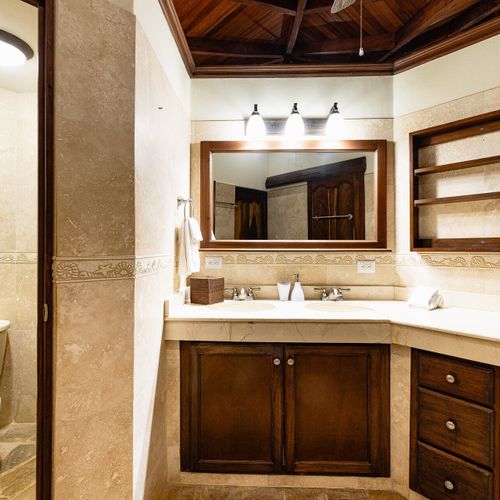 Travertine ensuite — double-sink hardwood vanity, vaulted wood ceiling, carved tile border.