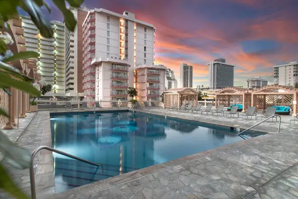 Shared swimming pool deck complete with poolside loungers, cabanas, and scenic Waikiki high-rise surroundings lit at dusk.