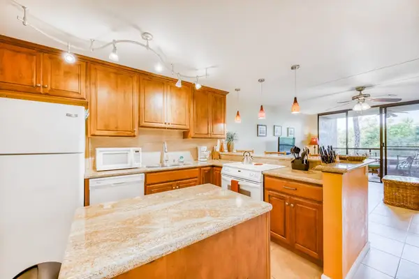 Spacious open kitchen featuring rich wooden cabinetry, a large prep island, white appliances, track lighting, and a view into the bright living room! 🎛️🪵📐