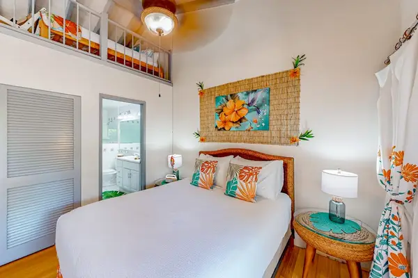 First bedroom presents queen bed, table, window, ceiling fan, artwork
