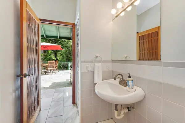 A seamless transition. This bright powder room pairs a classic pedestal sink and large mirror with a heavy wooden door opening directly onto the sunny outdoor patio. ☀️🚪