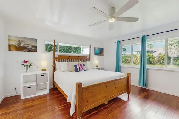 Coastal-style bedroom featuring a large wooden four-poster bed frame, warm hardwood floors, and vibrant teal curtains accenting wide windows that look out onto lush greenery.