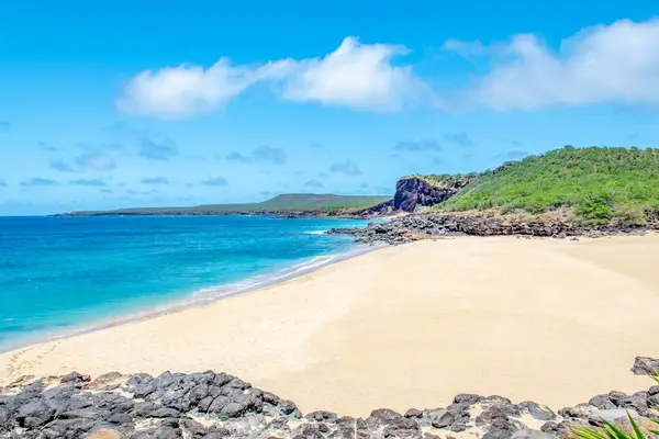 Sandy beach meets turquoise ocean beside rocky shoreline and hills