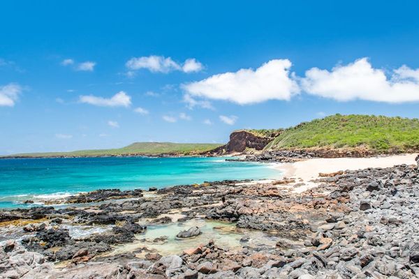 Rocky shoreline frames turquoise sea, distant headland, clouds, and sunlight