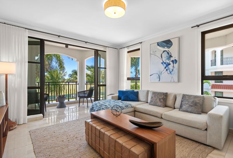 At Grand Reserve | Oceanfront Stay 3 This spacious, well-lit living room has a modern vibe with a comfy sectional sofa, coffee table, and large flat-screen TV. It's decorated in neutral colors with blue accents and has a sliding glass door that opens to a balcony with pool and greenery views. It’s a chill spot perfect for relaxing or hosting guests.