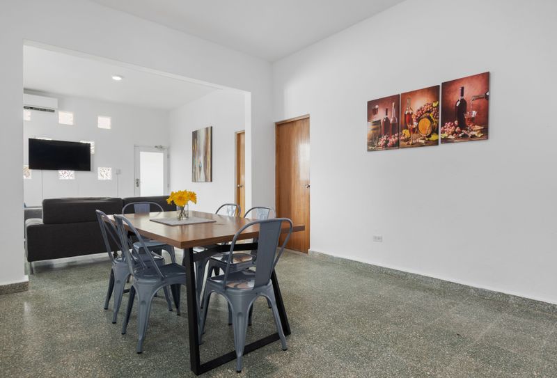 A modern dining room with a table that comfortably seats six or more people. Simple yet elegant design with cushioned chairs, a centerpiece, and plenty of space for group meals or gatherings.