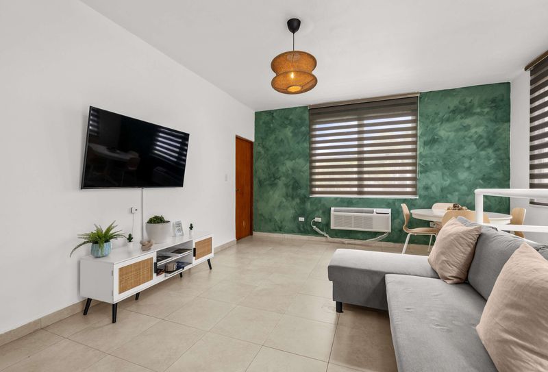 Cacique | Puerto Rico Vacation Rental 4 Sala con piso de baldosas claras. Televisor pantalla plana montado en pared blanca sobre consola baja con detalles de ratán. A la derecha, pared verde texturizada y parte de un sofá gris. Al fondo, comedor y ventana con persianas.