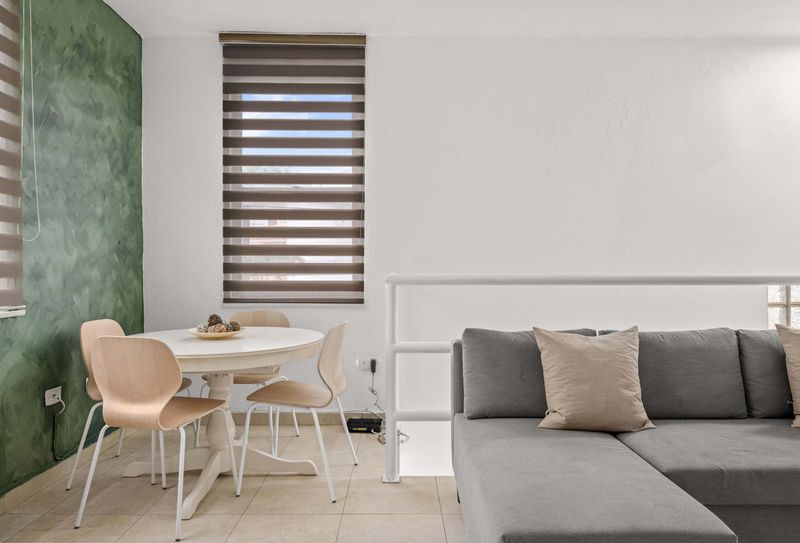 Cacique | Puerto Rico Vacation Rental 2 Espacio de concepto abierto. En primer plano, sofá seccional gris junto a una barandilla blanca. Al fondo, mesa de comedor redonda blanca frente a una pared verde. Dos ventanas con persianas y unidad de aire acondicionado instalada en la pared.