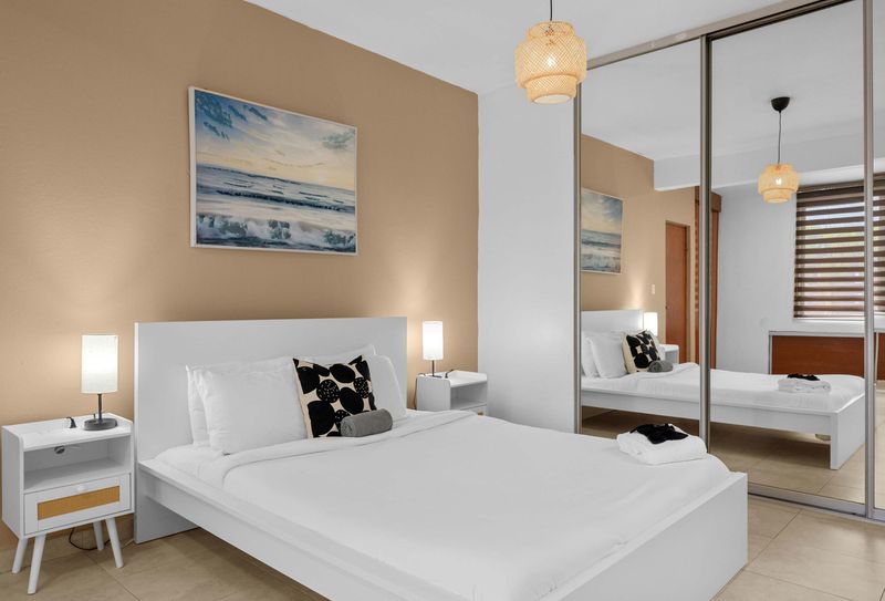 Cacique | Puerto Rico Vacation Rental 3 Dormitorio con cama blanca y pared beige con pintura de paisaje marino. A la derecha, un armario con puertas de espejo de piso a techo refleja la habitación y la ventana. Lámpara colgante de ratán en el techo y mesas de noche blancas a los lados.