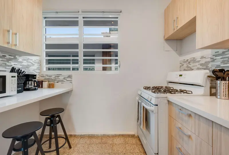 A narrow galley kitchen with light wood cabinets, a stainless steel gas stove, oven, and refrigerator. White countertops line both sides, with framed artwork on the wall and a wooden runner rug on the floor.
