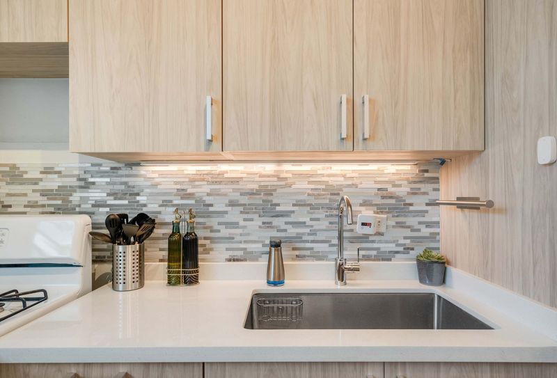 A narrow galley kitchen with light wood cabinets, a stainless steel gas stove, oven, and refrigerator. White countertops line both sides, with framed artwork on the wall and a wooden runner rug on the floor.