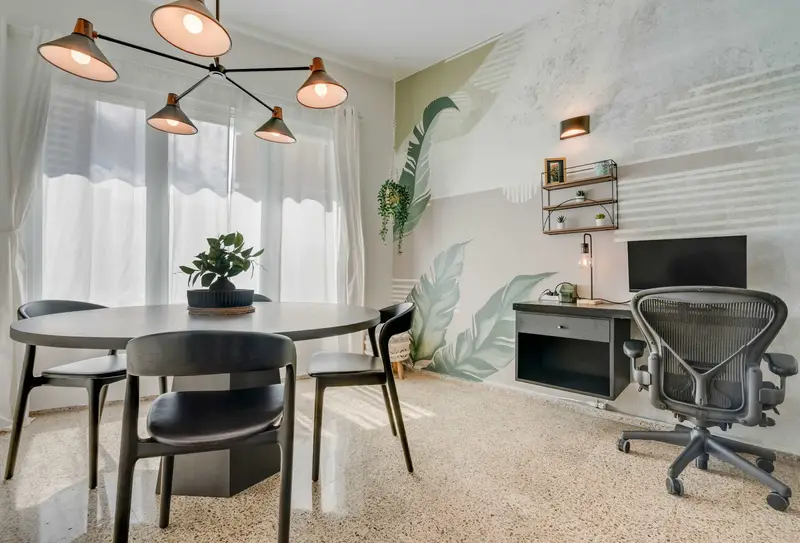 A dining area with a round black table and four matching chairs. Pendant lights hang above the table, and sheer white curtains cover the windows. Potted plants decorate the corners of the room. PC, desk and office chair