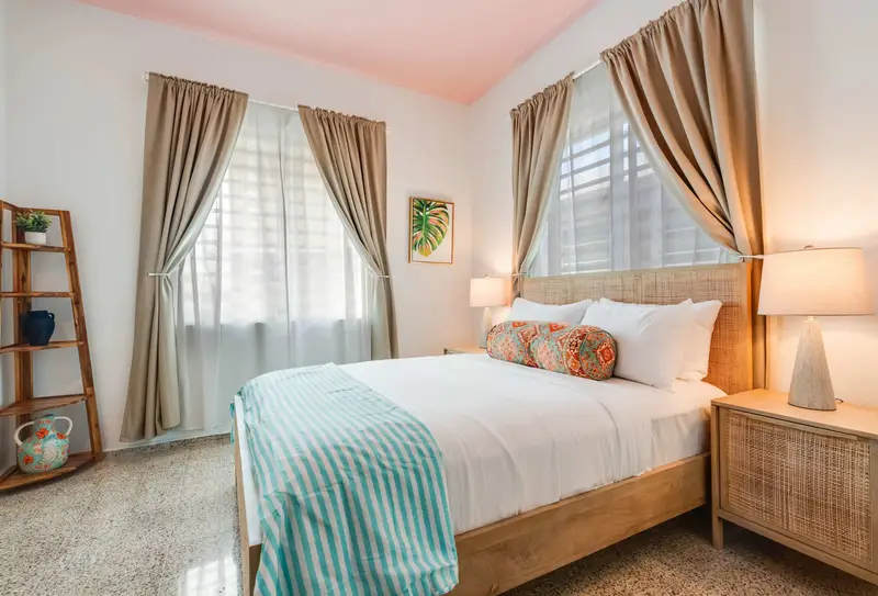A bedroom with a queen-size bed dressed in white linens and a light blue throw. Two windows with beige curtains allow natural light. A wooden nightstand and soft lighting frame the bed.