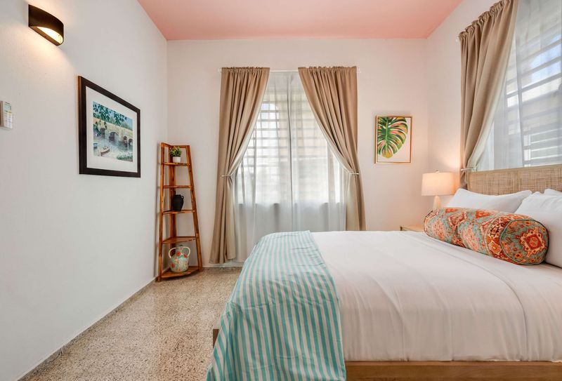 A bedroom with a queen-size bed dressed in white linens and a light blue throw. Two windows with beige curtains allow natural light. A wooden nightstand and soft lighting frame the bed.