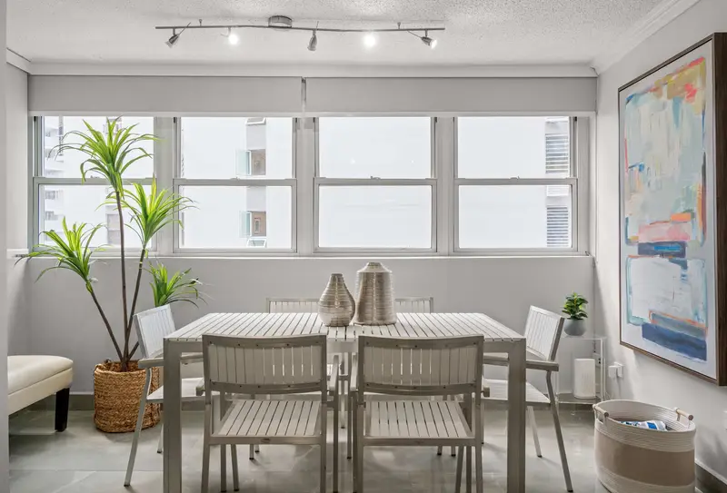 Walk to Beach | Free Parking | Condado 3BR 3 Second dining area with rectangular white table and six chairs beneath a row of windows with roller shades, potted palm and abstract art.