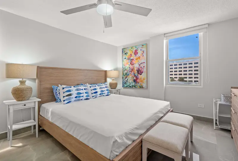 Walk to Beach | Free Parking | Condado 3BR 2 King bedroom with upholstered bench, woven-base lamps on white nightstands, colorful floral painting and window with city view.