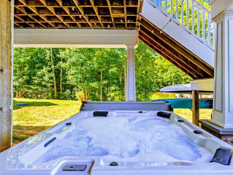 Hot Tub - A spacious hot tub tucked beneath the covered deck, bubbling and ready with green woods and water beyond.