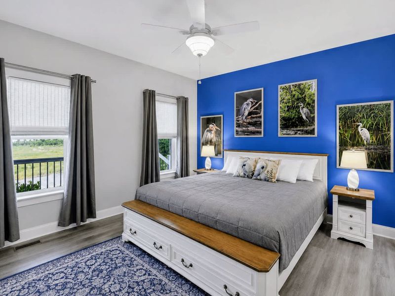 Primary Suite - King bed against a bold blue accent wall with heron photography and bright windows.