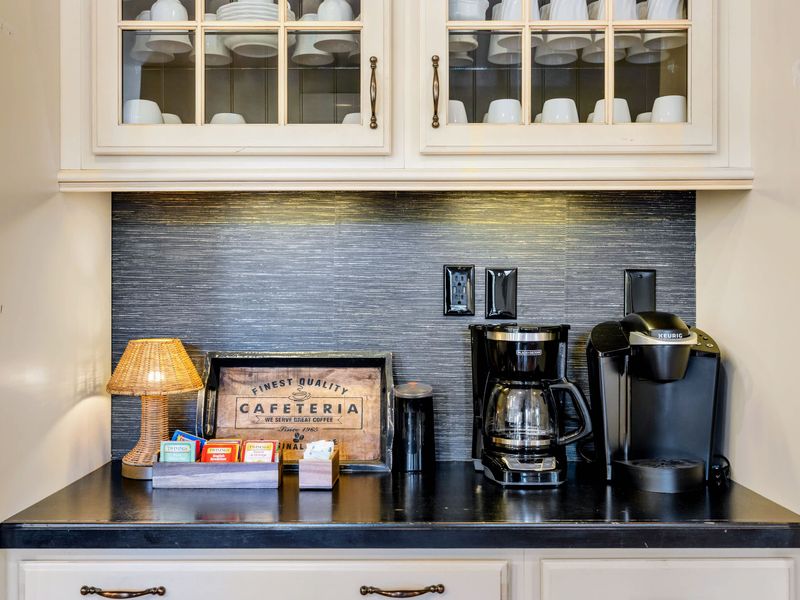 Kitchen - A dedicated coffee bar with drip brewer, Keurig, and a stocked tea caddy beneath glass-front cabinets of mugs.