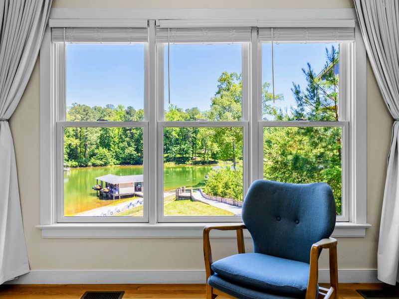 Secondary Suite - A blue armchair pulls up to triple windows overlooking the lake and a covered boathouse.