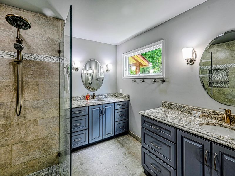 Primary Bath - A glass walk-in shower in warm stone tile beside a double granite vanity.