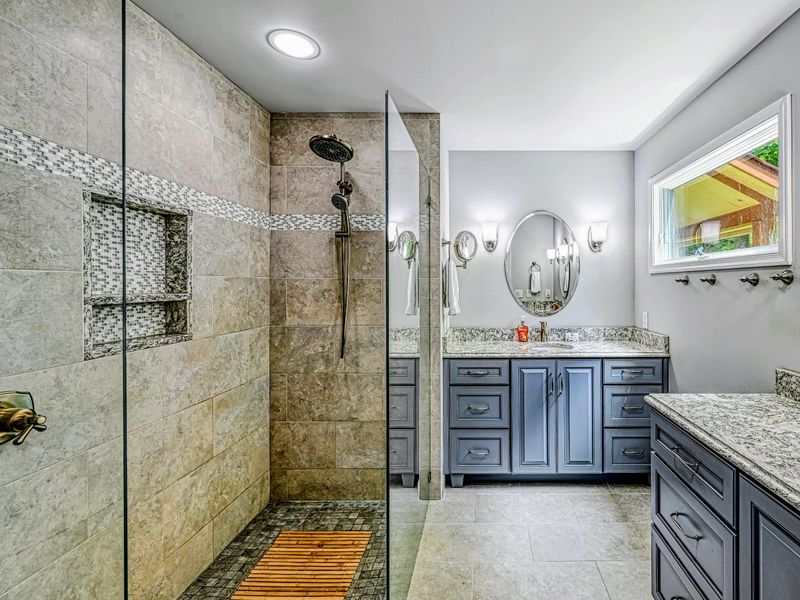 Primary Bath - A roomy walk-in shower and double granite vanities under soft sconce lighting.