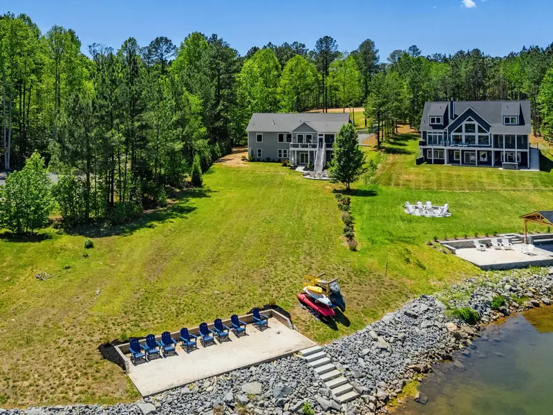 Shoreline Aerial - Blue Adirondack chairs line the lakeside patio with kayaks and a boat at the ready, lake life arranged and waiting.