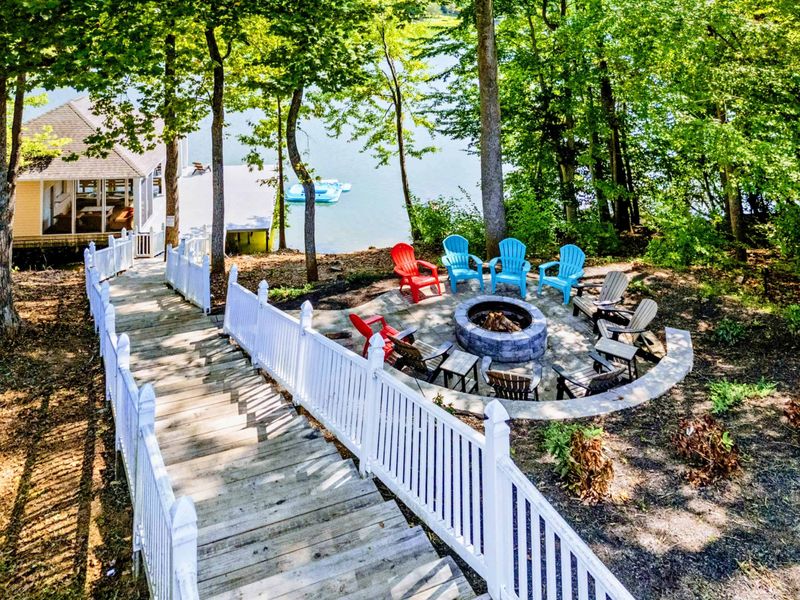 Fire Pit - The stone fire pit and colorful Adirondack chairs sit lakeside, framed by a white fence and the boathouse beyond.