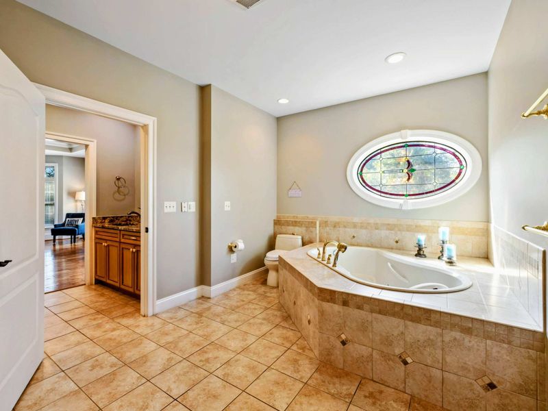 Primary Bath - A soaking tub beneath a stained glass oval window, with tile surround and gold fixtures.