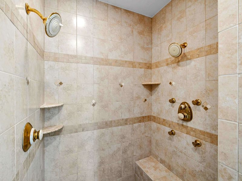 Primary Bath - A walk-in shower with dual gold showerheads, corner shelves, and tile trim details.