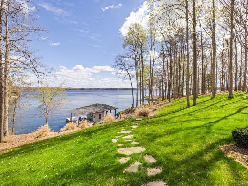 A serene lakeside view with a boat dock and a walking path leading through a grassy lawn flanked by a canvas of trees.