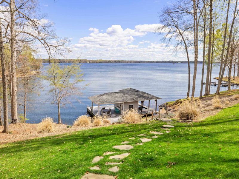 A serene lakeside view with a boat dock and a walking path leading through a grassy lawn flanked by a canvas of trees.