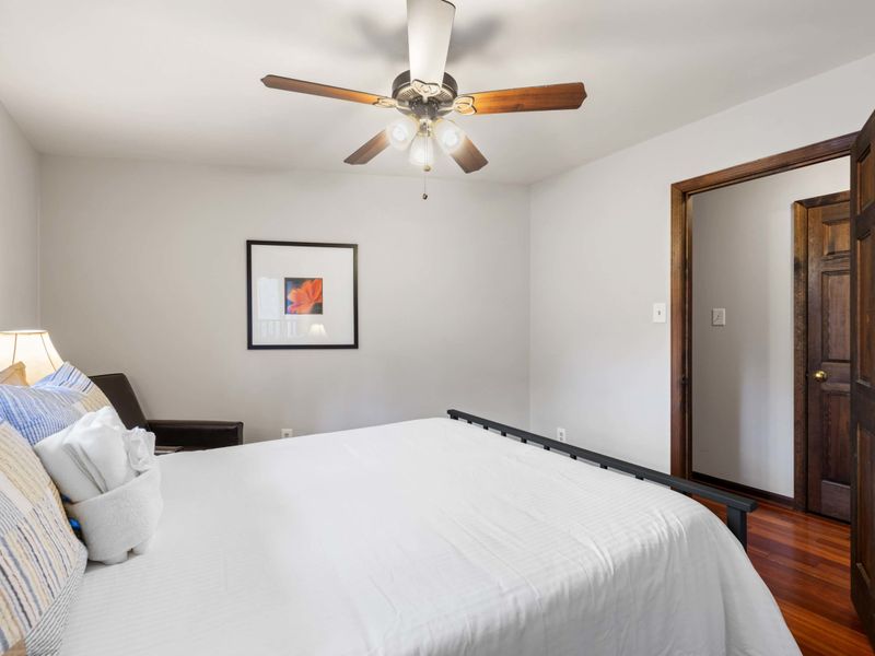 A simple and inviting bedroom with a ceiling fan, comfortable bedding, and warm wooden floors