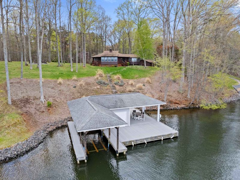 Overlooking a calm lake, a spacious covered dock sits at the water's edge with a pathway leading up to a secluded house