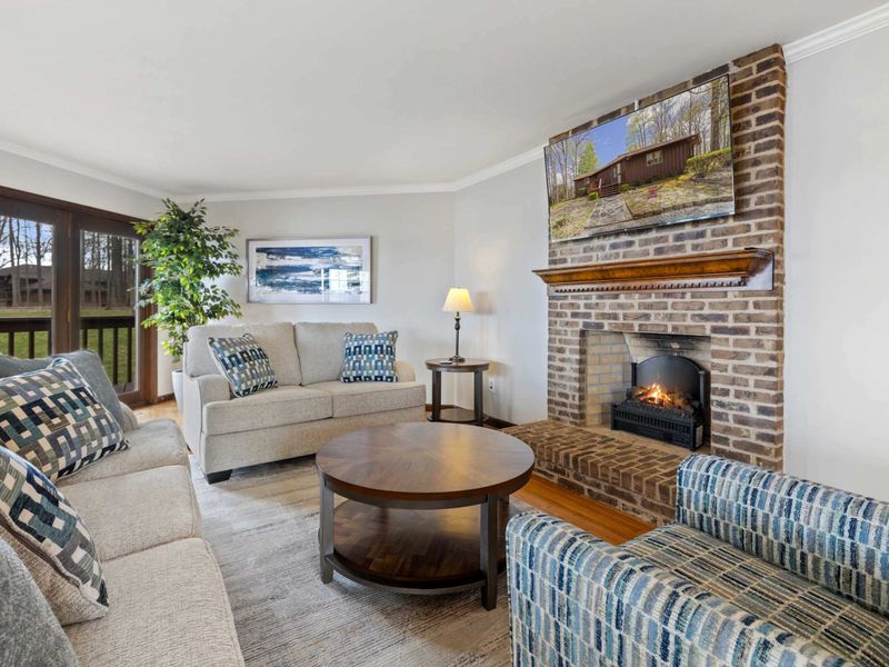 A warm and inviting living area with a classic brick fireplace and views of the lush outdoors through floor-to-ceiling windows