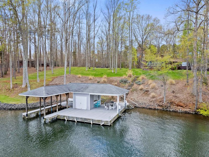 A tranquil lakeside scene featuring a covered boat dock with white chairs on the platform, surrounded by a forest of tall, thin trees and a well-kept grassy area leading to a house in the background.