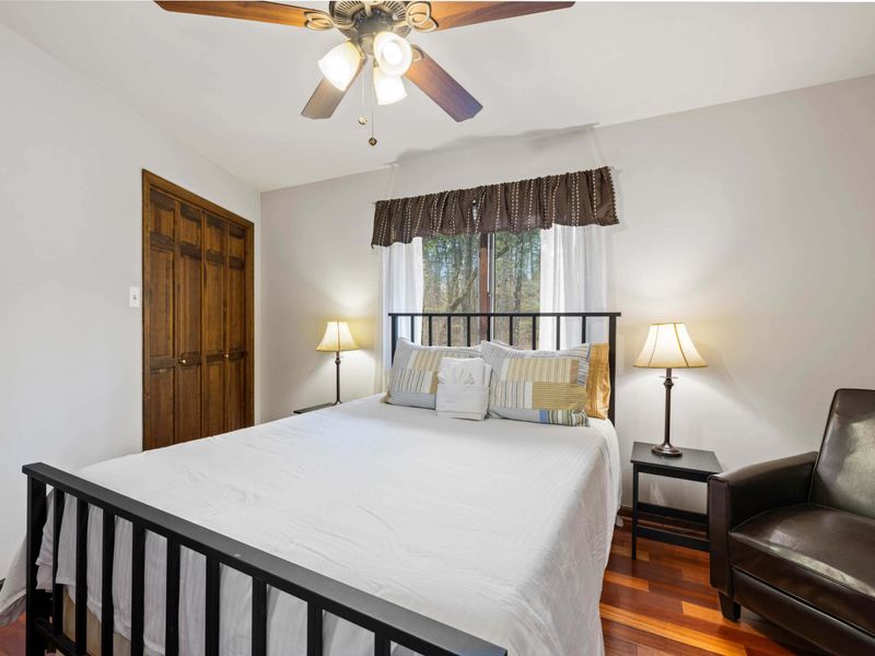 A simple and inviting bedroom with a ceiling fan, comfortable bedding, and warm wooden floors