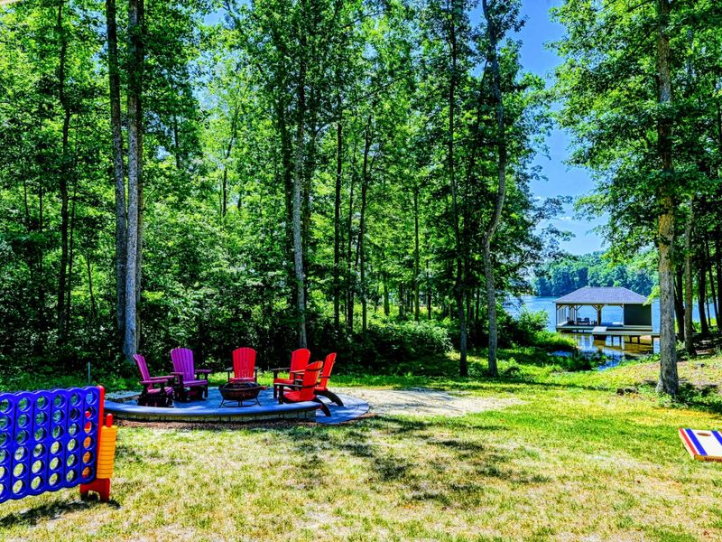 Spacious backyard with colorful Adirondack chairs around a fire pit, surrounded by trees and a view of the lake and dock.