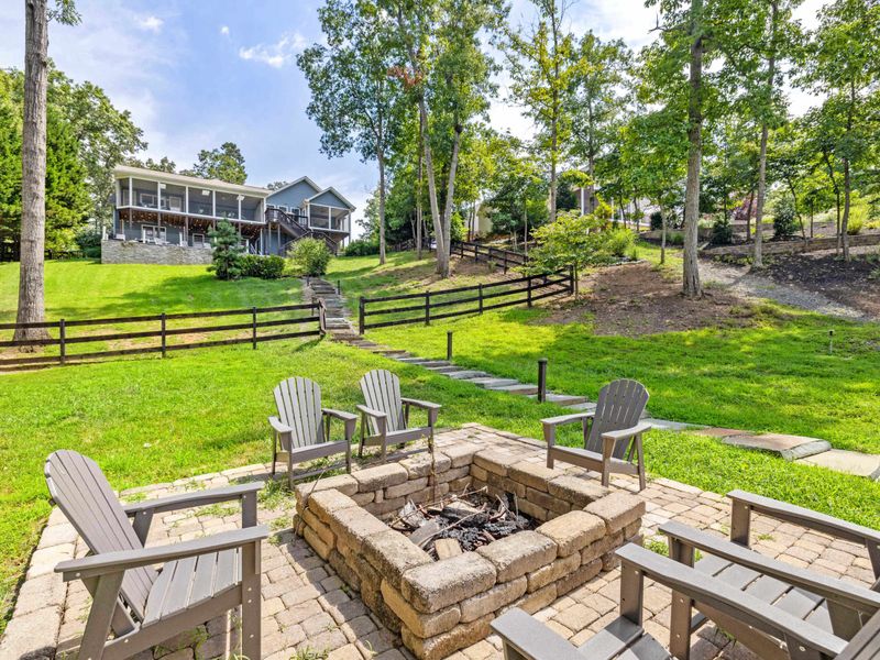 A cozy outdoor fire pit area surrounded by Adirondack chairs, set on a stone patio with lush greenery and easy access to the lake.