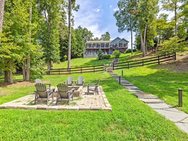 A cozy outdoor fire pit area surrounded by Adirondack chairs, set on a stone patio with lush greenery and easy access to the lake.