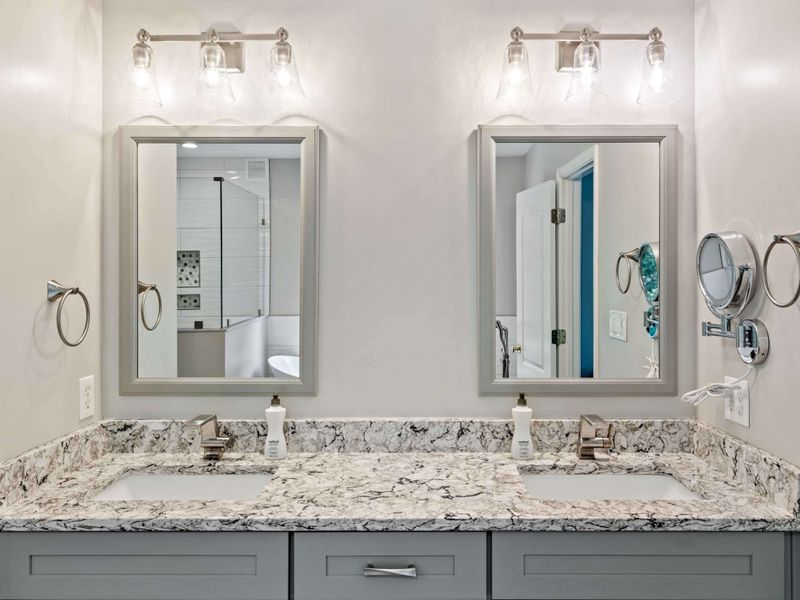 A bright and modern bathroom with a dual-sink vanity, granite countertops, and a view leading to the serene bedroom with lake vistas.